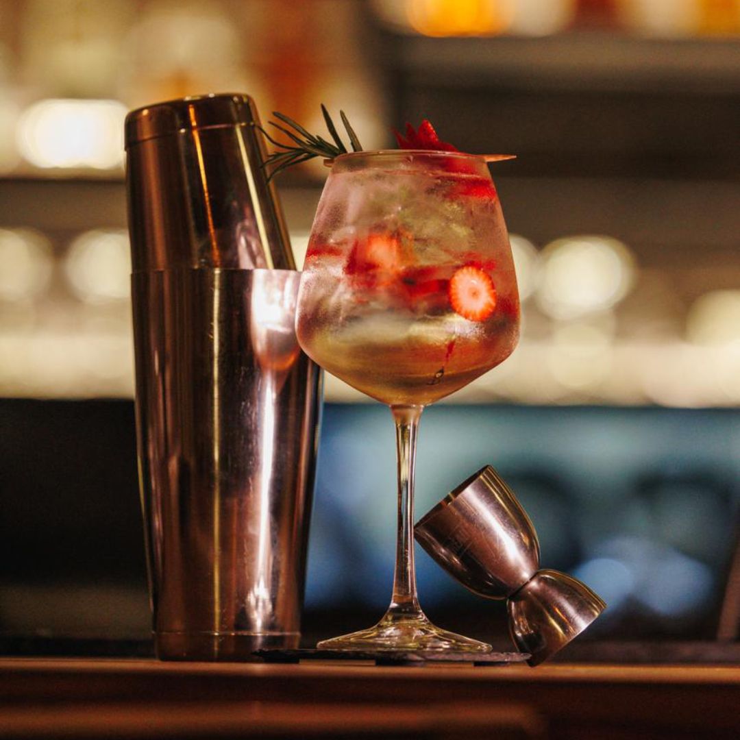 A stylish cocktail with raspberries beside a shaker on a bar counter.