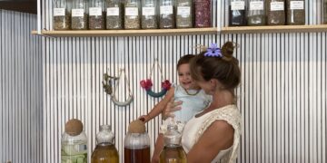 Yuli, clinical herbalist and owner of Marpe Jungle Apothecary, standing with her child at her shop counter surrounded by jars of dried herbs and teas in Santa Teresa Costa Rica