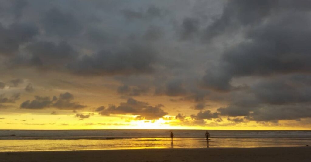 Two people standing on a beach at low tide as dark storm clouds reflect across wet sand during sunset.